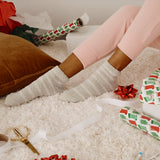 Person wearing fluffy socks on a carpeted floor with Christmas wrapping paper and ribbons.