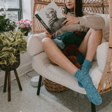 Person sitting on a white chair wearing fuzzy socks reading a book with plants and a wicker chair in the background.