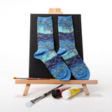A pair of crew socks with a blue and teal watercolor-style floral pattern, displayed on a black backdrop stand on a wooden artist easel with paint tubes and brushes.