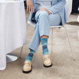 A person seated on a white chair wearing light blue tailored clothing and patterned blue socks with beige loafers, photographed in a real indoor setting with a table and floor visible. thumbnail