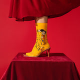 A person's leg wearing a bright yellow pointed-toe high-heeled boot with a surreal collage print, standing on a red velvet-covered pedestal against a solid red background.