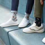 Two people standing on blue concrete steps, wearing patterned crew socks and sneakers. One person wears gray socks with baseball motifs and white sneakers, while the other wears black socks with football motifs and cream sneakers. thumbnail
