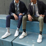 Two men sitting on blue concrete steps wearing white low-top sneakers and HOTSOX sports-themed novelty crew socks. They are dressed in casual jackets, shirts, and jeans or chinos. thumbnail