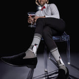 A man sits on a transparent chair wearing gray socks with penguin illustrations, black loafers, a white dress shirt, black trousers, and a red bow tie, while holding a martini glass against a black background. thumbnail
