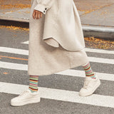 A person walking across a crosswalk wearing a long cream knit dress, a light beige cardigan, colorful striped socks, and white platform sneakers. thumbnail
