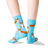 A pair of light blue crew socks with an all-over print of corgi dogs and small white bones, shown on a person's feet against a white background.