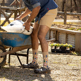 A person in a garden wearing beige crew socks with a colorful pine tree pattern, dark gray Crocs, and tan shorts, standing next to a blue wheelbarrow while holding a bag of soil or compost.