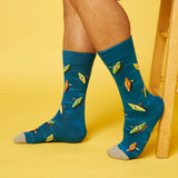 A person's legs wearing blue crew socks with a pattern of cartoon kayaks, standing against a solid yellow background next to a wooden stool.