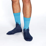 A person's legs wearing bright blue crew socks with a dark navy blue dripping graphic design on the feet, photographed against a seamless white background.