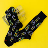 A pair of black novelty socks with embroidered beer bottles and beer mugs, displayed on a bright yellow background next to a clear glass mug. thumbnail