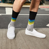 A person wearing gray crew socks with a black top band and multicolor chevron stripes in blue, green, and yellow, paired with white slip-on shoes on a city sidewalk. thumbnail