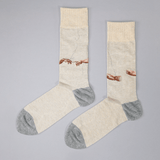 A pair of crew socks with a beige and gray color-blocked design, featuring a printed graphic of two hands reaching toward a crack in the wall, styled on a neutral gray background.