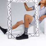A person in light gray pajamas sits behind a large decorative white frame, wearing black slip-on shoes and a gray crew sock with a black-and-white face graphic. The photo is taken in a clean white studio setting. thumbnail