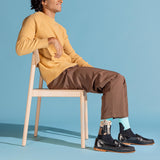 A man sits on a light wood chair wearing a mustard yellow ribbed sweater, brown trousers, black loafers, and patterned socks against a solid light blue studio background. thumbnail