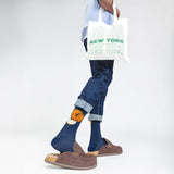 Person wearing blue jeans and brown shoes with a white tote bag featuring 'New York' text. thumbnail