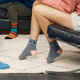 A person sitting on a black chair wearing gray socks with a cat face design and pink toes, with another person's legs visible in the background wearing colorful striped socks in a home setting. thumbnail