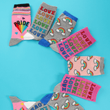 Six pairs of colorful novelty socks arranged in a circle on a bright turquoise background. The socks feature various designs including rainbow clouds, the word 'LOVE' repeated, 'PRIDE' with a rainbow, and blue cuffs.