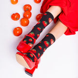 Person wearing black socks with red tomato pattern and red shoes, surrounded by tomatoes on a light background thumbnail