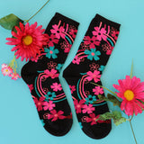 A pair of black crew socks decorated with vibrant pink, teal, and orange floral patterns, arranged on a light blue background with decorative pink flowers. thumbnail
