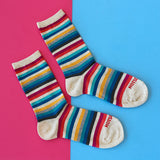 Three crew socks with multicolor horizontal stripes in red, orange, yellow, teal, blue, black, and white, laid out on a split pink and blue background. The toes are cream-colored, and the word 'HOTSOCKS' is printed in red on the toes. thumbnail