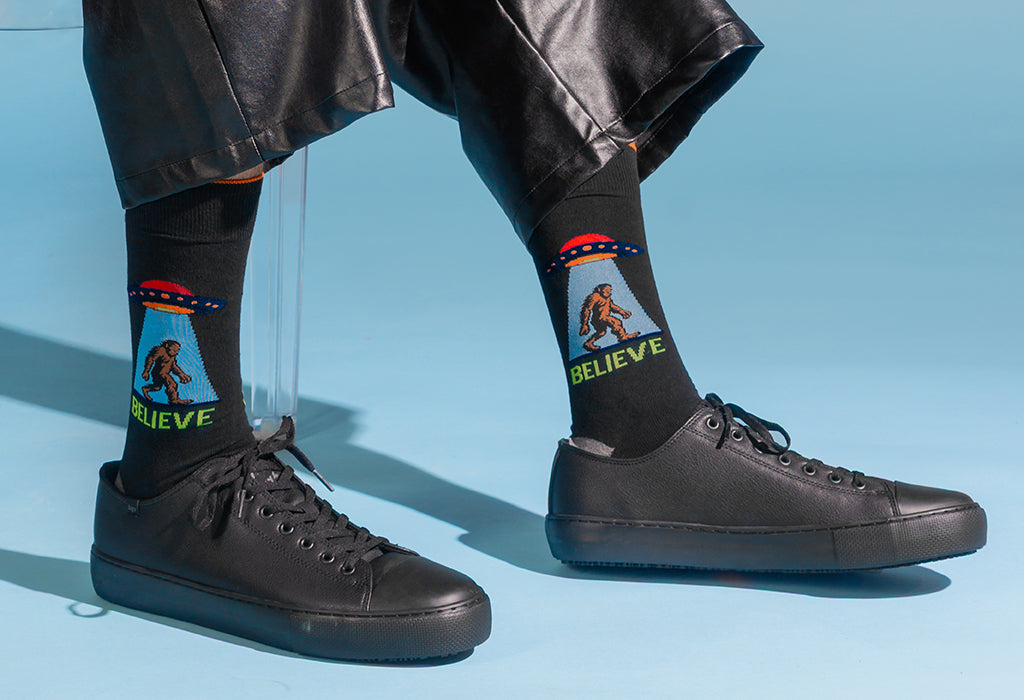 Black crew socks with a graphic of a Bigfoot-like figure under a UFO and the word 'BELIEVE' in green, worn with black sneakers and black pants against a light blue background.