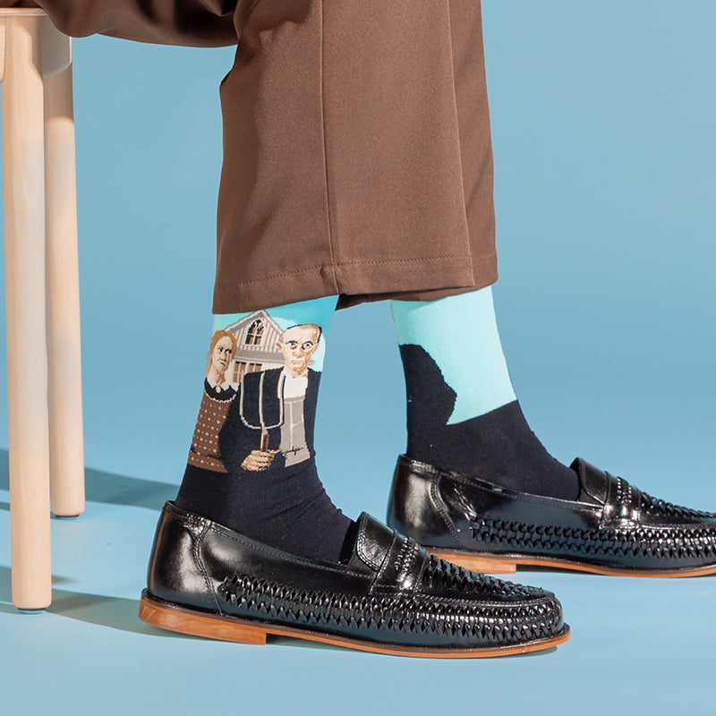 A person seated on a stool wearing black woven loafers, brown trousers, and dark socks with a colorful illustrated print on one leg and a light blue section on the other, against a light blue studio background.