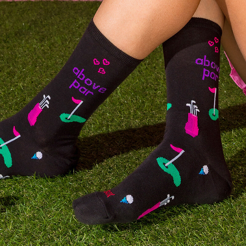 A pair of black crew socks with a colorful golf-themed print and the text 'above par' worn on feet on green grass.