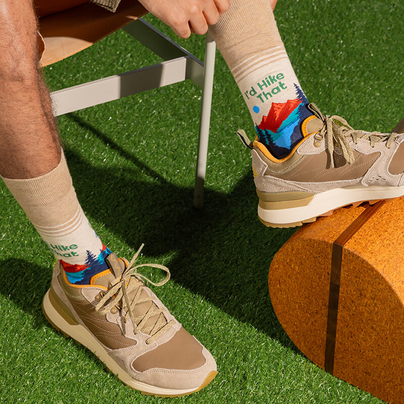 A person seated outdoors on artificial grass wearing beige hiking socks with a colorful mountain graphic and the phrase “I'd Hike That,” paired with tan and beige athletic sneakers.