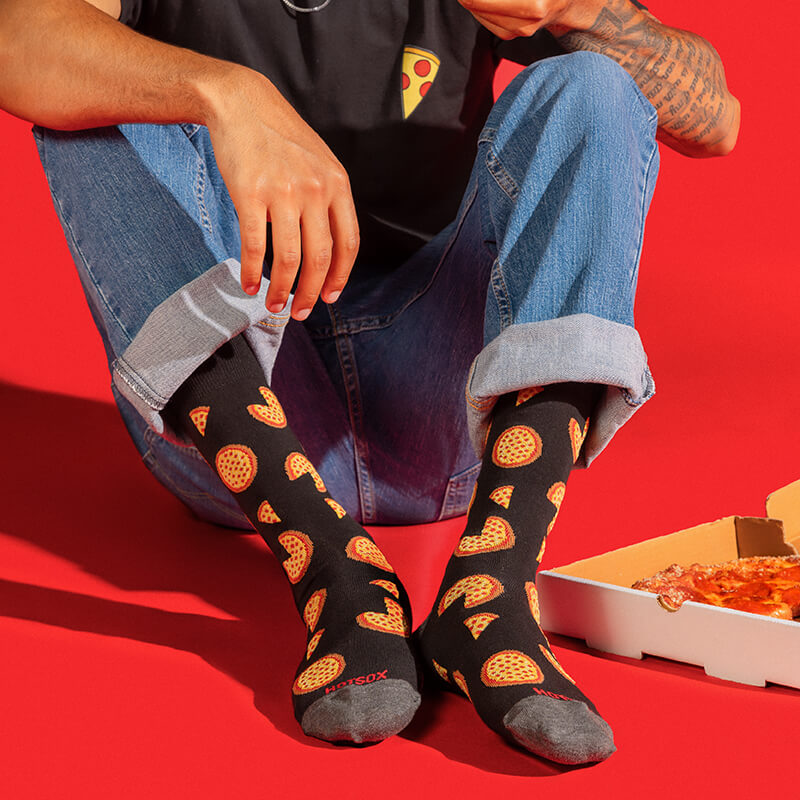 A person sitting on a red floor wearing black socks with a repeating pizza slice pattern, blue jeans, and a black graphic t-shirt. A pizza box is visible on the right side.