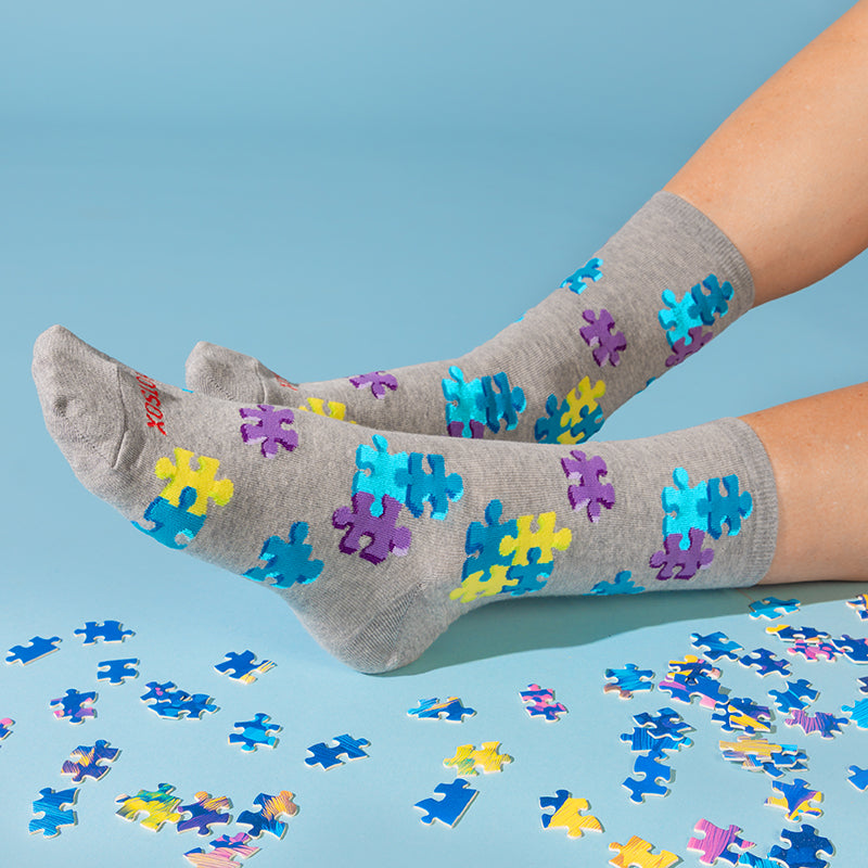 Gray crew socks with colorful puzzle-piece patterns in blue, purple, and yellow, worn on feet with scattered loose puzzle pieces on a light blue background.