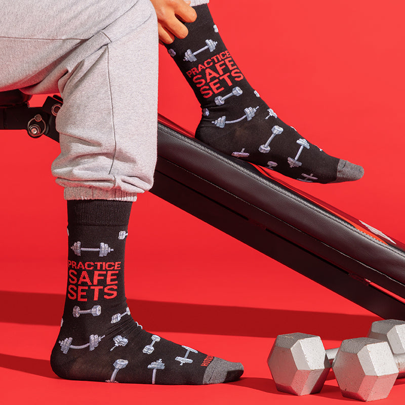 A person wearing black crew socks with red and white gym-themed text and dumbbell illustrations, seated on a red bench with dumbbells nearby against a bright red background.