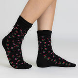 A pair of black crew socks with a small pink and green floral pattern, shown on a model's feet against a light gray studio background. thumbnail