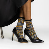 A person seated in a black chair wearing black pointed-toe high heels and metallic-striped crew socks with black ruffled tops. The socks have horizontal stripes in black, mustard yellow, gray, and white.