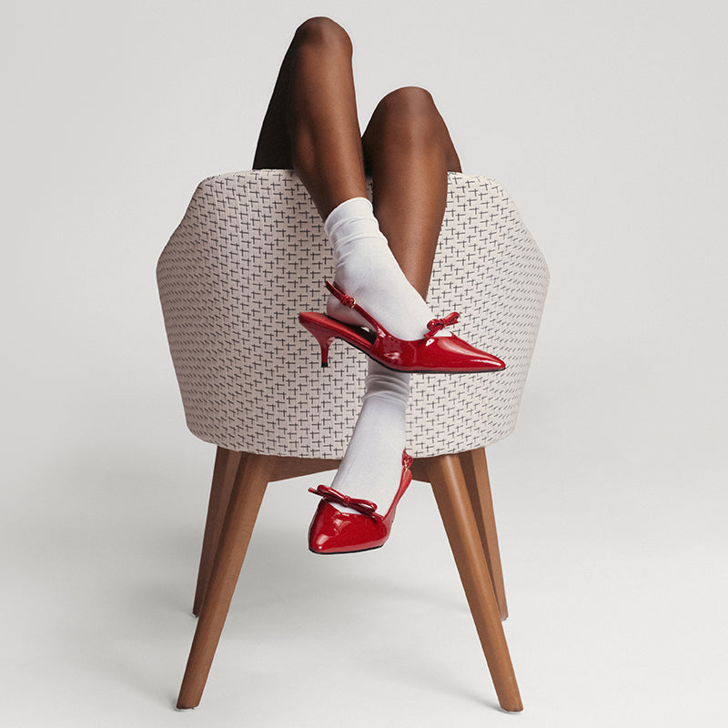 A person seated in a woven chair with their legs crossed, wearing white socks and shiny red slingback heels against a light gray studio background.