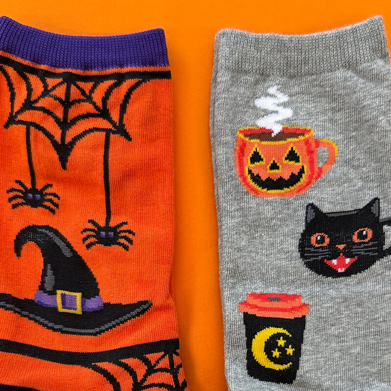 Two pairs of knit socks laid flat on an orange background. The left sock is bright orange with a purple cuff and Halloween graphics including black spiders, a witch hat, and spiderwebs.