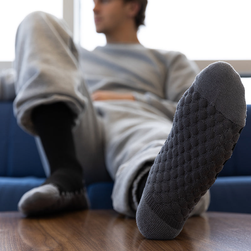 A person reclining on a blue couch with their legs stretched, showcasing a dark gray knit slipper sock with a textured diamond pattern on the sole, while wearing a black slipper on the other foot.