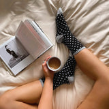 Woman holding a coffee cup, sitting on a bed wearing HOTSOX black and white polka dot socks while reading a book. thumbnail