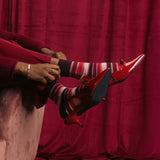 Person wearing red high-heeled shoes and striped socks sitting on a pink couch against a red curtain.