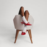 Person wearing red high-heeled shoes and white socks sitting on a woven stool against a plain background