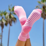 Women's pink and white striped hydrating K.Bell crew socks with aloe in front of palm trees and a clear blue sky. thumbnail