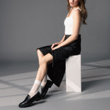 Woman sitting on a white block wearing a black skirt, white top, and black shoes with white striped socks. thumbnail
